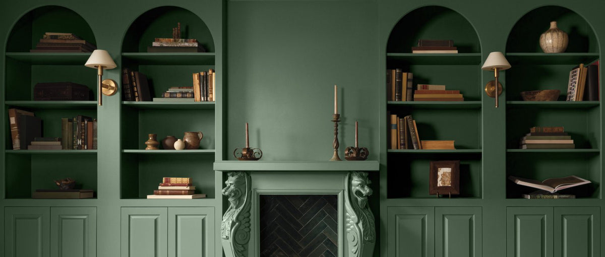A green painted room with lower paneled walls and arched, built-in shelving topped with books and decor.